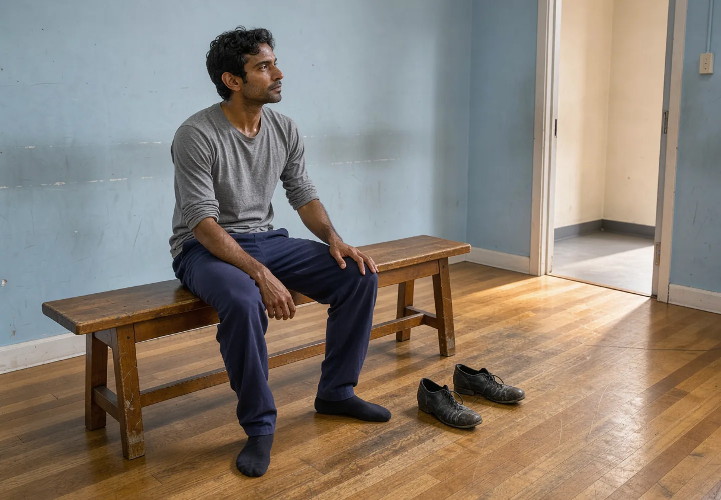 A dancer rests by an open studio doorway, with rehearsal shoes beside the bench.